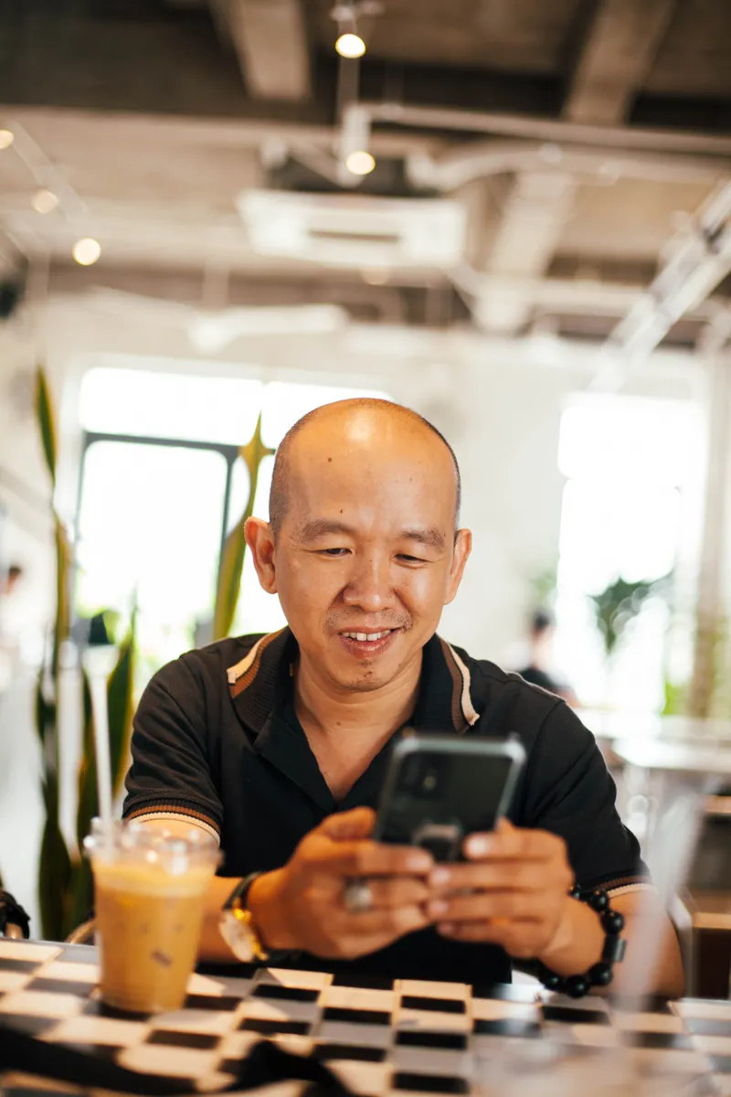 A smiling man relaxes in a bright, airy space, reading his phone with an iced coffee in hand.