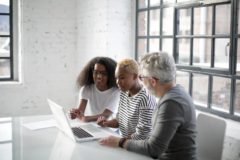 Three colleagues of different ages gather around a laptop at a white table in a bright office with tall windows.