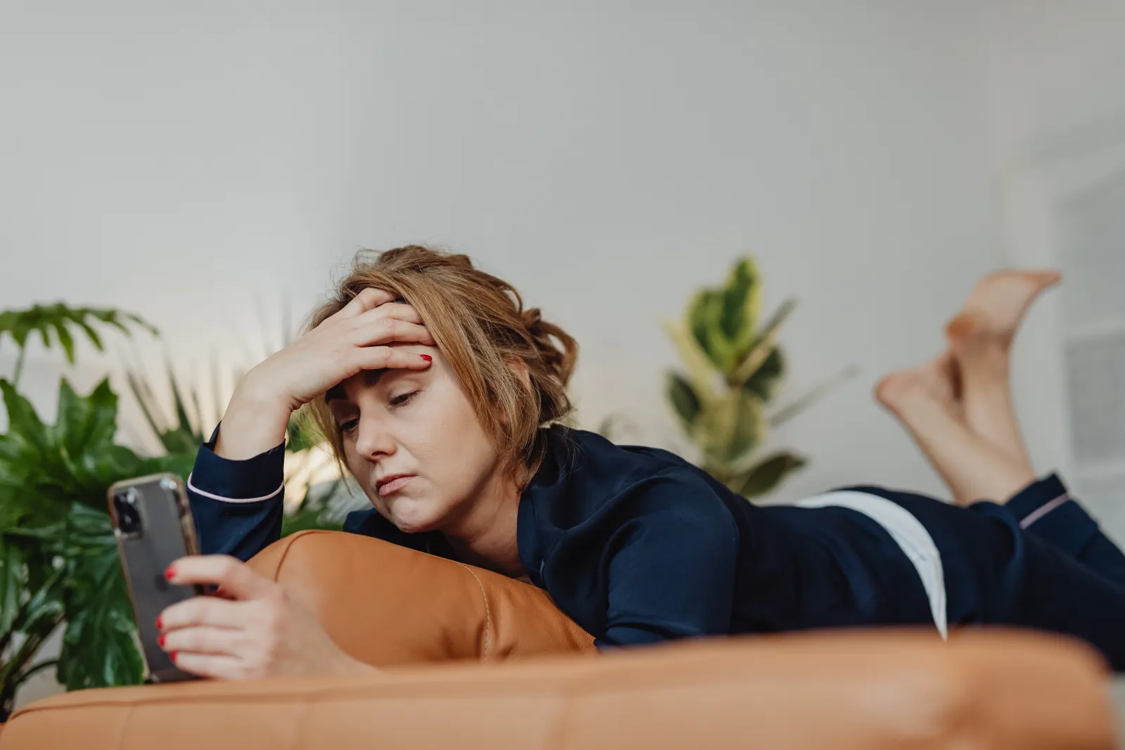 A woman lies on an orange couch in a bright room with plants, one hand pressed to her forehead, frowning at a text on her phone.